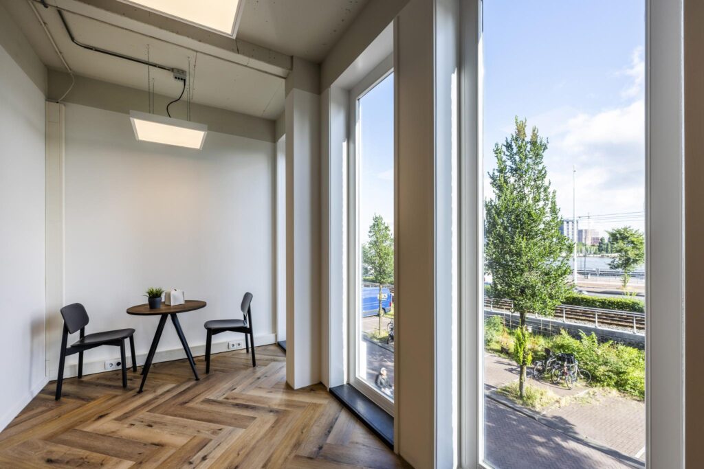 Zithoek met twee stoelen en een rond tafeltje bij hoge ramen met uitzicht op de De Ruijterkade in Amsterdam.